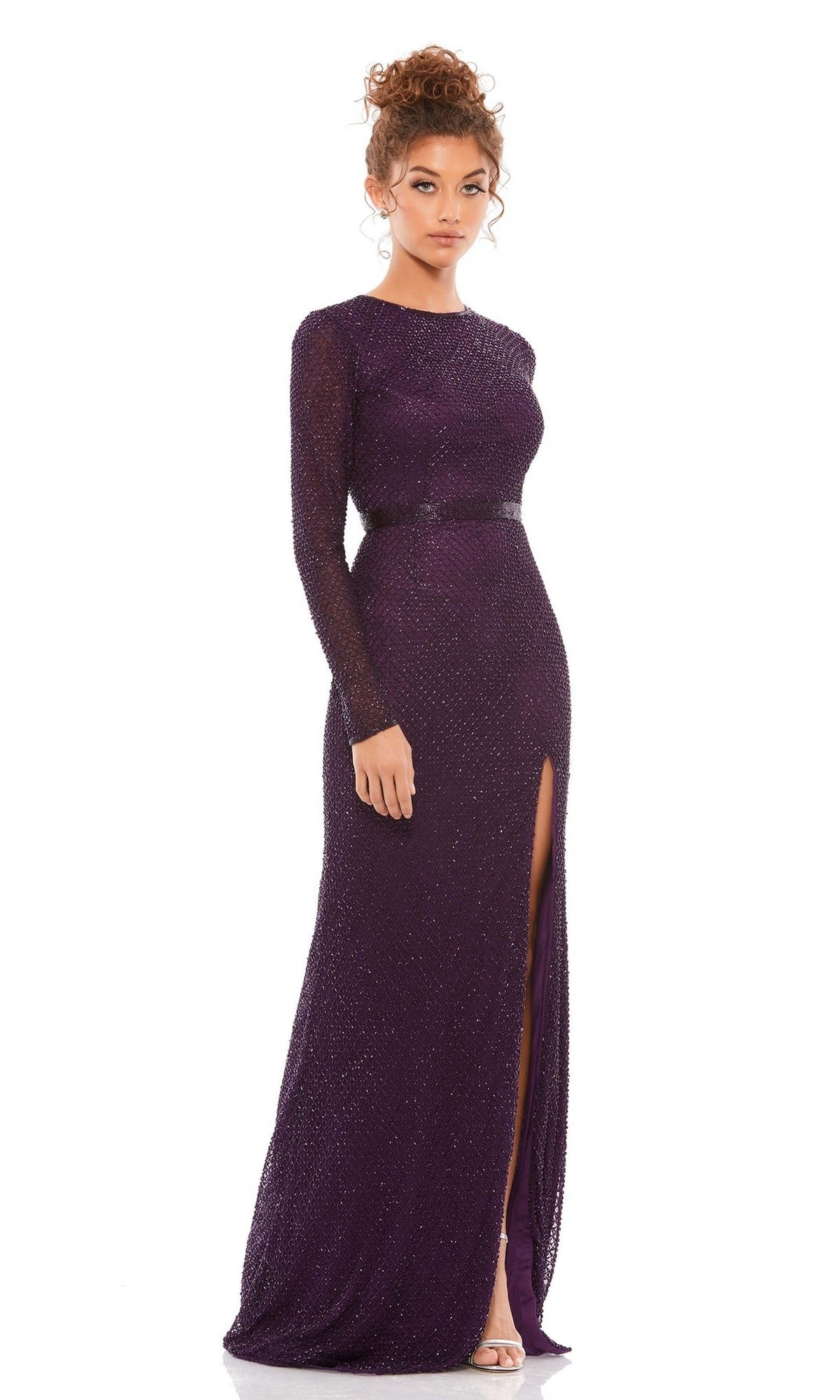 A woman in the Long Formal Dress 5056 by Mac Duggal, featuring fitted long sleeves, a sparkly dark purple fabric, and an elegant slit, stands against a white background.